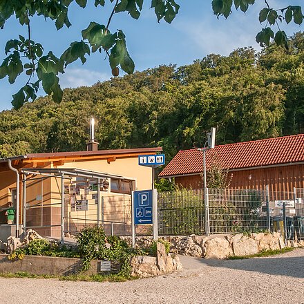Das Gebäude aus Holz ist l-förmig gebaut und schön mit Pflanzen und Steinen angepflanzt. Hinter dem Campingplatz sieht man den Wald. Der Himmel strahlt himmelblau.