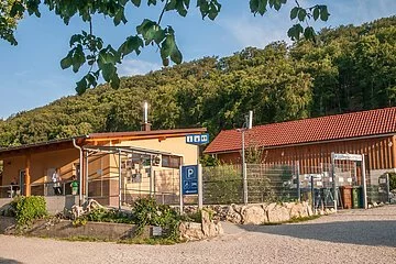 Das Gebäude aus Holz ist l-förmig gebaut und schön mit Pflanzen und Steinen angepflanzt. Hinter dem Campingplatz sieht man den Wald. Der Himmel strahlt himmelblau.