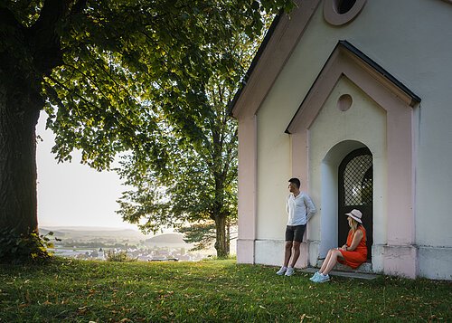 Ein Mann im weißen Shirt lehnt an der Wand einer Kapelle an, eine Frau im roten Kleid und Strohhut sitzt auf der Stufe. Beide genießen den Sonnenuntergang. Links neben der Kapelle steht ein großer Baum mit grünem Blätterdach, dass den Beiden Schatten spendet.