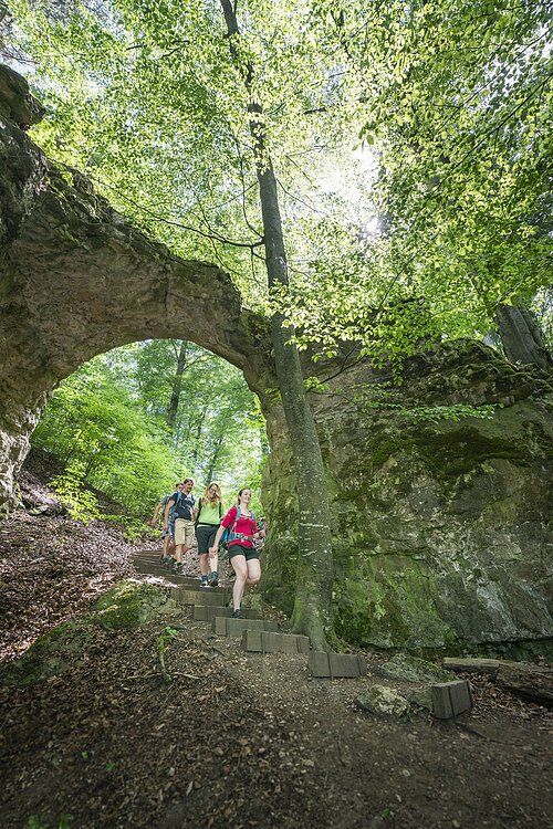 Eine kleine Wandergruppe steigt die Stufen durch das Felsentor hinunter. Hinter dem Tor erstreckt sich der Wald, wo die Sonne ein wenig durchblitzt.