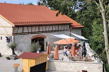 Vor einer renovierten Säule stehen Tische und Stühle auf einer sonnigen Terrasse, die von einer Steinmauer eingefasst ist.