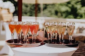 Auf einem weißen Partystechtisch befinden sich zwei braune Tabletts. Auf einem Tablet stehen Weinbläser mit weißem Sekt und auf dem anderen Weingläser mit rose farbenem Sekt.