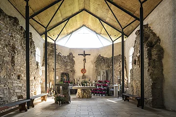 Innenraum einer Kapelle mit Altar, Kreuz, Kerzen und Blumen, umgeben von alten Steinmauern und Holzdecke
