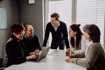 Fünf Personen sitzen und stehen an einem Tisch, eine Frau arbeitet an einem Laptop, alle wirken im Gespräch vertieft.