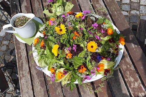 Bunter Salat mit essbaren Blüten auf weißem Teller neben weißem Krug mit Kräuterdressing auf Holztisch.