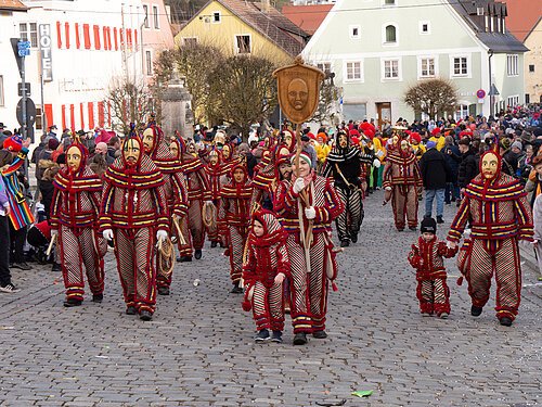 Menschen in gestreiften Kostümen und Holzmasken bei Umzug auf gepflasterter Straße vor Zuschauern