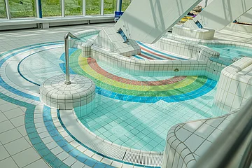 Innenbereich eines Schwimmbads mit kleinem Becken und bunten Fliesen in Regenbogenfarben am Boden.