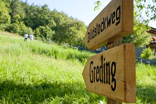 Holzwegweiser mit den Aufschriften „Bildstockweg“ und „Greding“ vor einer grünen Wiese und zwei Personen im Hintergrund