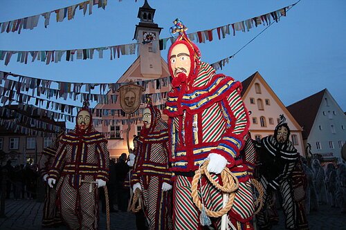 Menschen in gestreiften Kostümen und Masken bei einem abendlichen Fest mit Girlanden und historischen Gebäuden.