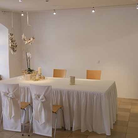 Raum mit großem Tisch, zwei Stühlen mit weißen Hussen und Deko, Fenster links, Pflanze rechts, weiße Wände. Dieser Saal dient als Trausaal.