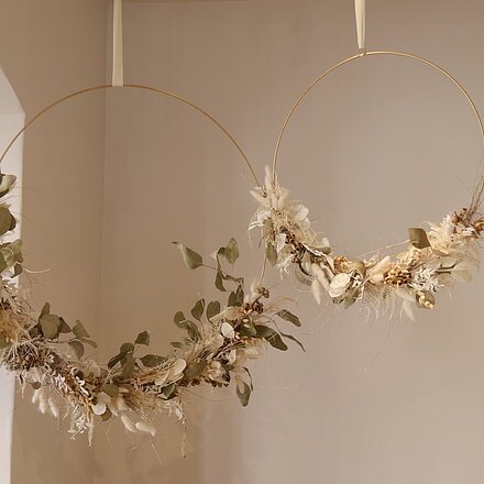Zwei hängende goldene Metallringe mit getrockneten Blättern und Blumen an weißen Wänden.