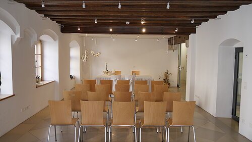 Trausaal mit Holzstühlen in Reihen, weißer Wand, Holzbalkendecke und Tisch mit weißer Tischdecke vorne.