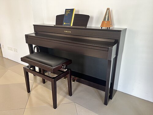 Braunes Yamaha-Klavier mit Klavierbank, Buch und Metronom auf Fliesenboden vor weißer Wand.