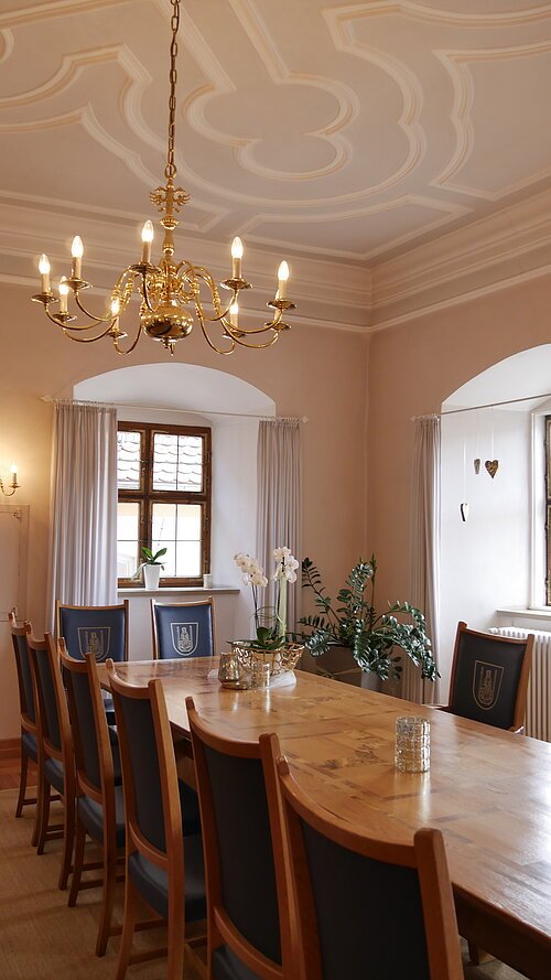 Standesamt Trauzimmer im Gredinger Rathaus mit langem Holztisch, Holzstühlen mit blauen Polstern, Kronleuchter und Fenstern.