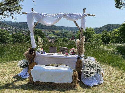 Traualtar auf dem Kalvarienberg mit weißer Dekoration, Blumen und Stühlen vor grüner Wiesen- und Berglandschaft.