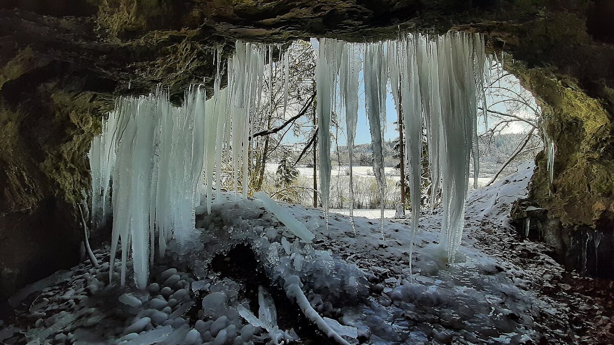 Blick aus einer Höhle mit großen Eiszapfen an der Decke auf eine verschneite Winterlandschaft mit Bäumen.