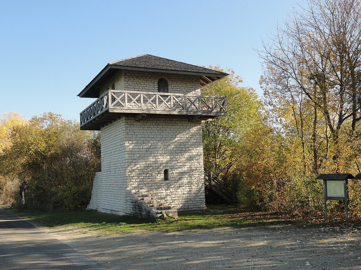 Steinerner Aussichtsturm mit Holzbalkon und Treppe, umgeben von herbstlichen Bäumen neben einem Weg.