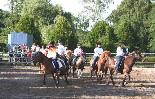 Reiter in weißer Kleidung und Helm reiten auf Pferden in einem Außenreitplatz vor Zuschauern.