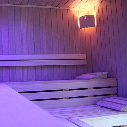 Sanarium Innenraum einer Sauna mit Holzbänken, Wanduhr und indirekter Beleuchtung in violettem Licht