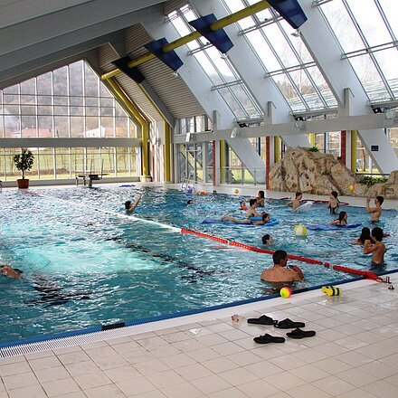 Innenaufnahme eines Schwimmbads mit Schwimmern und spielenden Personen im Wasser, große Fensterfront sichtbar.