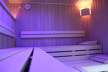 Innenraum einer Sauna mit Holzbänken, Wanduhr und indirekter Beleuchtung in violettem Licht
