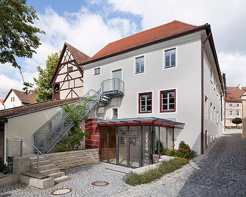 Eingang einer Bücherei in einem weißen Gebäude mit rotem Dach und Außentreppe aus Metall, gepflasterter Straße.