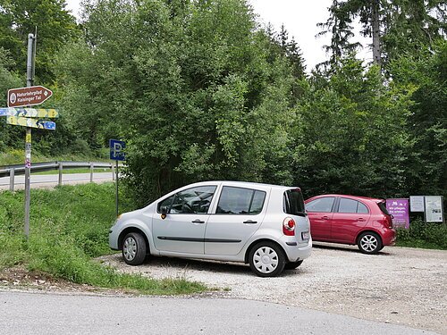 Parkplatz mit zwei Autos vor Wald und mehreren Wegweisern, darunter „Naturlehrpfad Kaisinger Tal“
