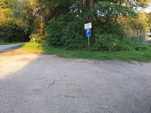 Parkplatzschild am Waldrand neben unbefestigtem Weg und angrenzendem Asphaltweg im Sonnenlicht