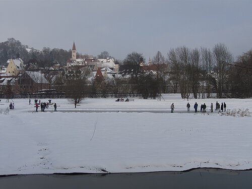 Schneebedeckte Wiese mit Menschen vor einem Dorf mit verschneiten Dächern und kahlen Bäumen im Winter.