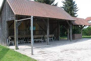 Überdachter Holzpavillon mit Tischen und Bänken auf gepflastertem Boden neben Rasenfläche und Bäumen.
