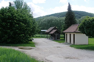 Freier Platz mit zwei kleinen Gebäuden und Bäumen vor bewaldetem Hügel unter bewölktem Himmel