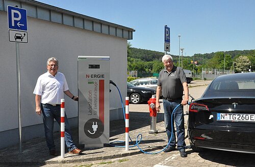 Zwei Männer an Ladestation für Elektrofahrzeuge, ein Mann hält Ladekabel an schwarzem Tesla, Parkplatz mit E-Ladeschild.