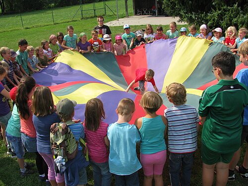 Kinder stehen im Kreis und halten ein großes, buntes Tuch auf einer Wiese, ein Kind klettert darauf.