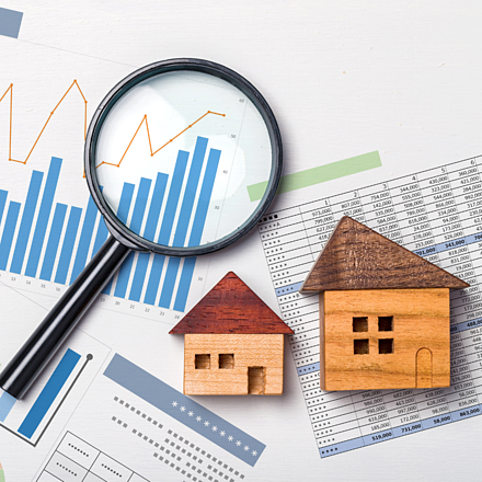 Bodenrichtwerte Lupe über Diagrammen und Tabellen neben zwei kleinen Holzhausmodellen auf weißem Untergrund.