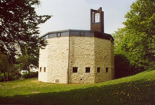 Runde Kirche aus hellem Stein mit kleinem Glockenturm und Kreuz, umgeben von Bäumen und Wiese.
