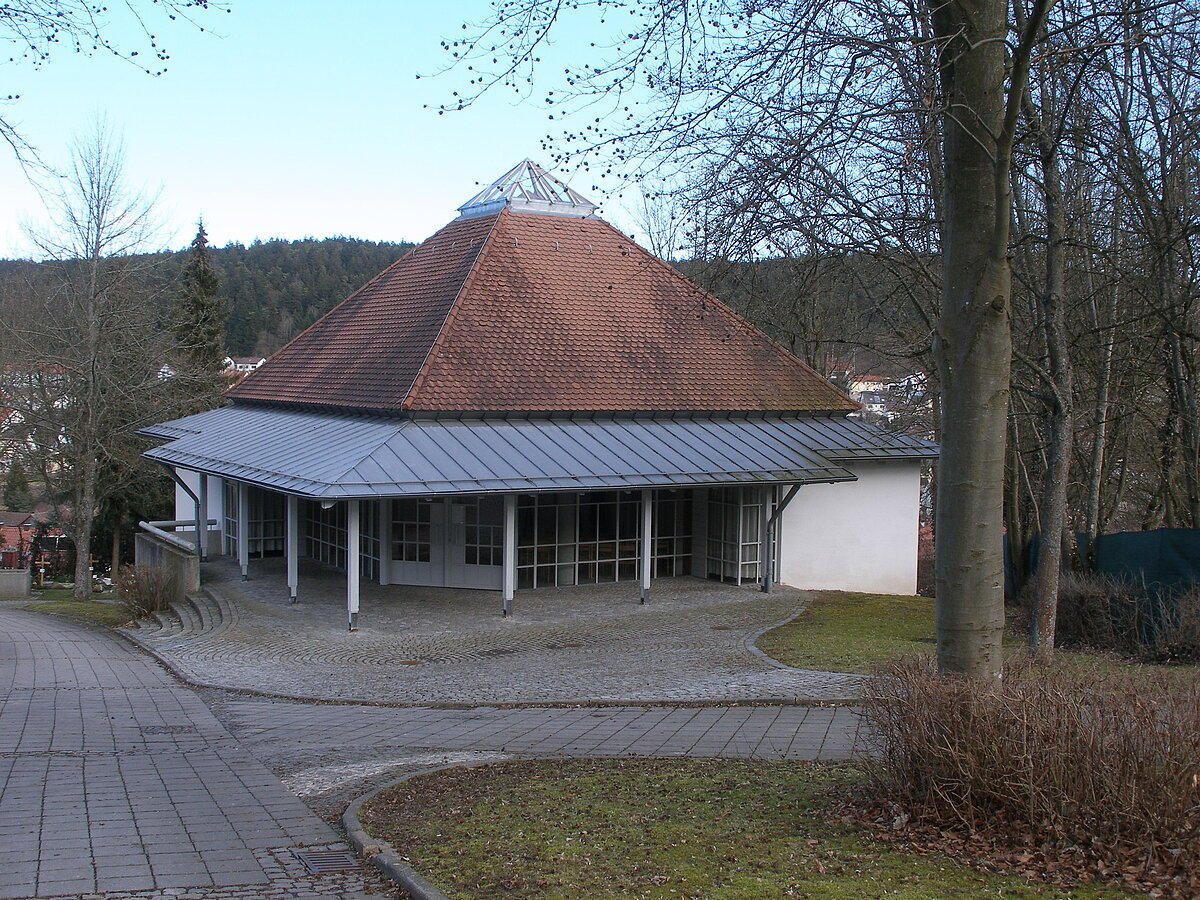 Gebäude mit pyramidenförmigem Dach und umlaufendem Vordach, umgeben von kahlen Bäumen und Gehwegen.