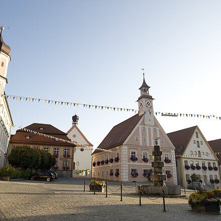 Marktplatz von Greding Historische Gebäude mit Turm und Blumenschmuck an einem gepflasterten Platz mit Girlanden bei klarem Himmel.