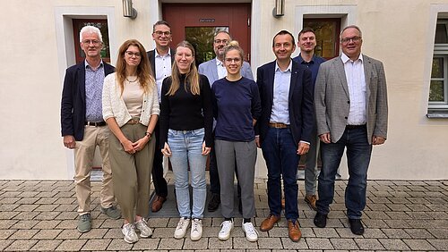 Neun Personen stehen vor einem Gebäude mit zwei Türen und zwei Fenstern auf gepflastertem Boden.