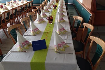 Langer Tisch mit weißer Tischdecke, grüner Tischläufer, gefalteten Servietten und Blumenarrangements, Stühle drumherum.