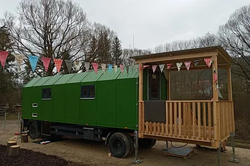 Grüner Wohnwagen mit Holzveranda und bunten Wimpeln, umgeben von kahlen Bäumen an bewölktem Tag.