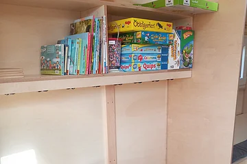 Regal mit Kinderbüchern und Gesellschaftsspielen in einem hellen Holzraum mit Telefon und Teppichboden.