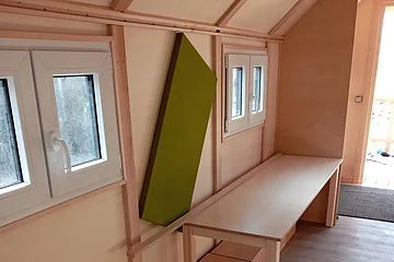 Innenraum mit Holzwänden, zwei kleinen Fenstern, einem langen Tisch und mehreren quadratischen Holzkisten als Sitzgelegenheiten.