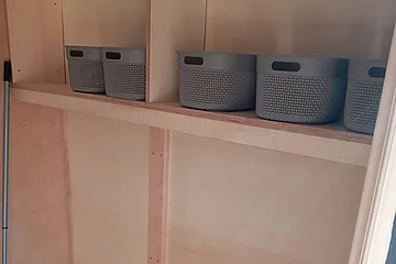 Holzregal mit grauen Kunststoffkörben und zwei Holzbänken in einem kleinen Raum mit Besen.