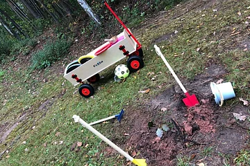 Kinderwagen mit Spielzeug, Schaufel, Rechen und Eimer auf Grasfläche mit Wald im Hintergrund