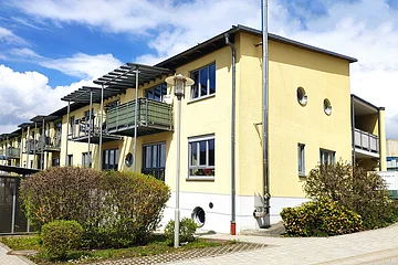 Mehrfamilienhaus mit gelber Fassade, Balkonen, runden Fenstern und Büschen bei sonnigem Wetter.