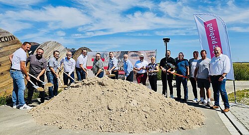 Gruppe von 15 Personen mit Schaufeln vor großem Sandhaufen bei Baustellenbeginn im Freien bei Sonnenschein.