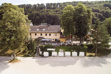 Hotelgebäude mit Terrasse und Sonnenschirmen umgeben von Bäumen vor bewaldetem Hügel im Hintergrund.