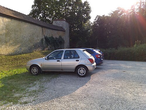 Drei parkende Autos auf einem Kiesplatz neben einer alten Steinmauer und Bäumen bei Sonnenschein.