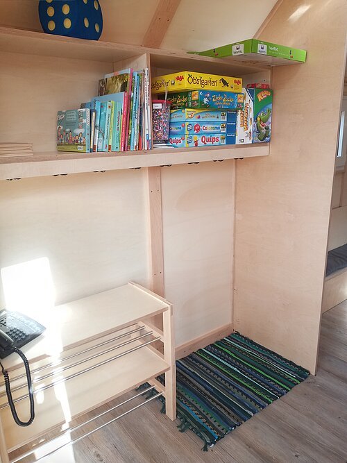 Regal mit Kinderbüchern und Gesellschaftsspielen in einem hellen Holzraum mit Telefon und Teppichboden.
