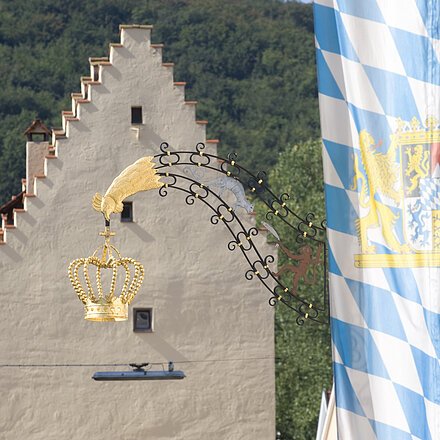 Goldene Krone an geschwungenem Metallarm vor einem Haus mit Treppengiebel und bayerischer Flagge im Hintergrund